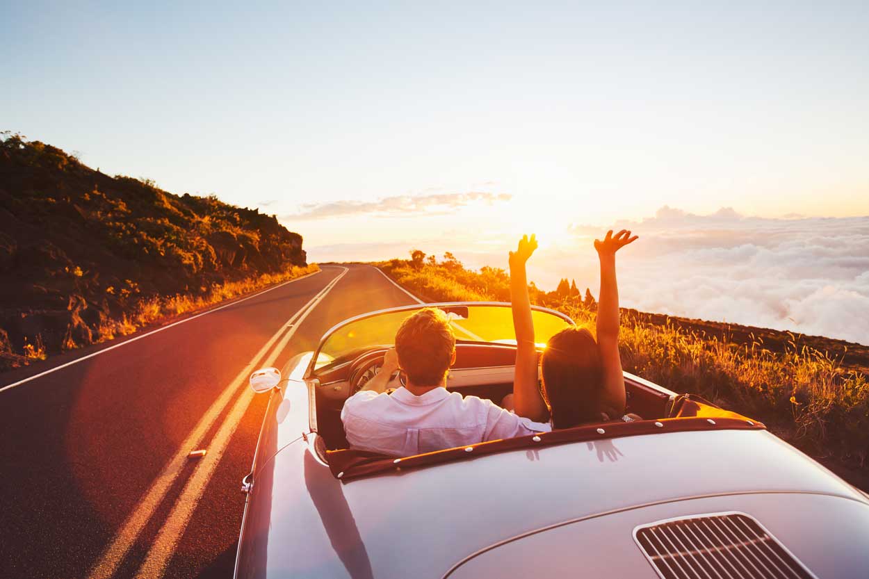 How to plan a road trip, two people in a white convertible car, driving down a hilly road towards the sunset.