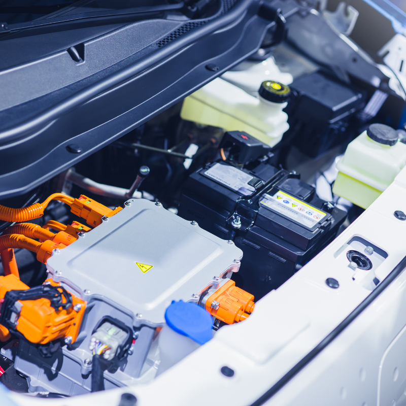Close-up view of an electric vehicle's engine compartment, highlighting orange wiring, a silver control unit with a yellow warning symbol, and various fluid reservoirs.